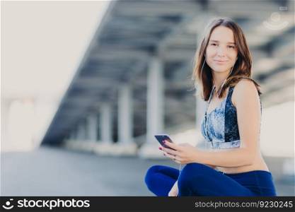 Pleased woman with healthy skin, feels refreshed while listens favourite track in earphones, dressed in sportswear, sits in lotus pose, waits for friend, poses outdoor. People and lifestyle concept