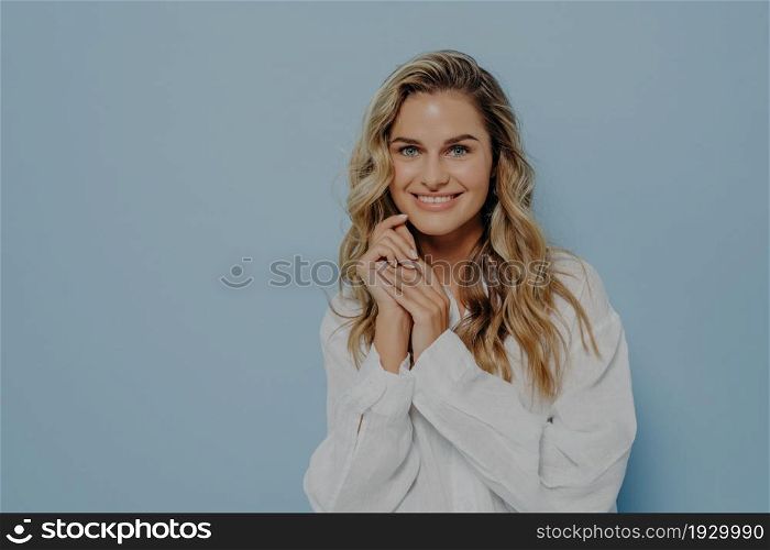 Pleased young caucasian woman with blonde wavy hair wearing white casual shirt smiling broadly and keeping hands near face, beautiful female glad to hear good positive news. Happy people concept. Pleased young caucasian woman with blonde wavy hair wearing white casual shirt smiling broadly at camera