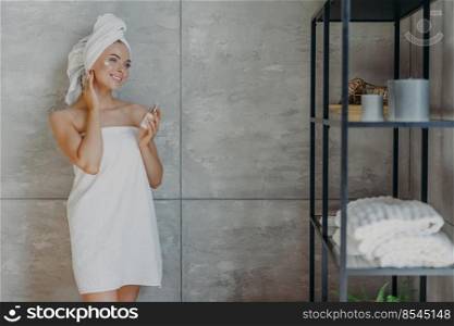 Pleased young European woman applies face lotion holds bottle of cosmetic product wrapped in white bath towel stands against grey wall in bathroom. People cosmetology beauty and cleansing concept