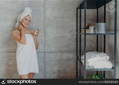Pleased young European woman cares of her beautiful skin, applies moisturizer on face, holds bottle of lotion, stands against grey wall in bathroom, wrapped in bath towel, has refreshed look