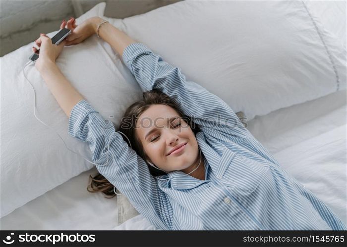 Pleased young European woman stretches in bed, has closed eyes, dressed in striped nightclothes, holds smart phone, enjoys favourite music in earphones, lies on white bedclothes in cozy bedroom