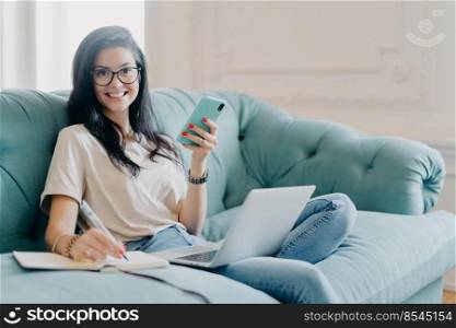 Pleased young female designer with dark hair creats new project, holds modern smartphone in hands, makes notes in notepad, poses on sofa, uses laptop computer connected to wireless internet.