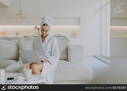 Pleased young female model concentrated at laptop screen wears bathrobe and towel on head poses in spacious living room at sofa enjoys spending weekend at home feels comfortable and relaxed.