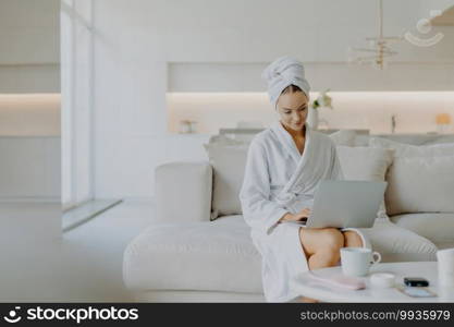 Pleased young female model concentrated at laptop screen wears bathrobe and towel on head poses in spacious living room at sofa enjoys spending weekend at home feels comfortable and relaxed.