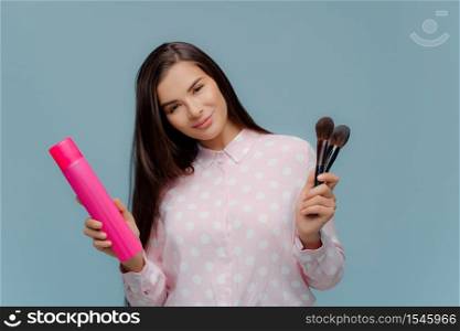 Pleased young female model makes hairstyle and makeup, holds bottle of hairspray and cosmetic brushes, tilts head, dressed in elegant clothes, poses over blue background. Beauty and style concept