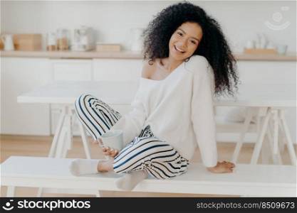 Pleased young girlfriend with Afro curly hairstyle, sits crossed legs on bench, enjoys morning coffee and domestic atmosphere, poses against kitchen background, spends time alone, has breakfast