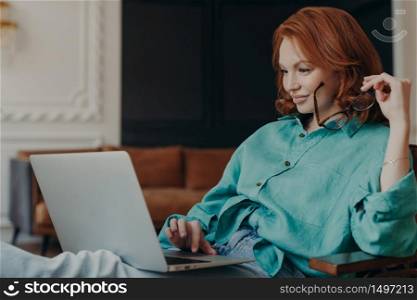 Pleased young redhead woman sits on comfortable armchair at home, does remote work, reads news in social networks, busy with computer job, concentrated in monitor of laptop. Modern technologies