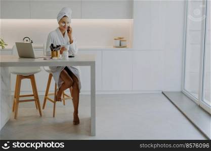Pleased young woman dressed in bathrobe poses bare feet at chair in spacious kitchen sits at table with laptop and cosmetic products applies beauty patches under eyes talks via modern cellular