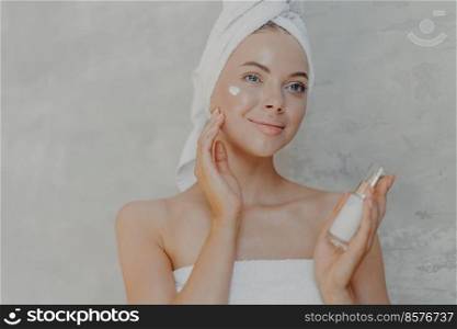 Pleased young woman has healthy glowing skin, minimal makeup, applies face lotion and looks happily into distance, wears wrapped towel on head and around body, poses against grey background.