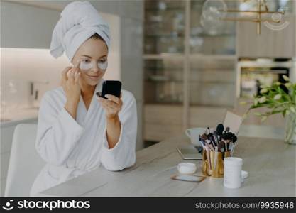 Pleased young woman wears bathrobe and towel wrapped on head cares for under eye skin applies beauty patches looks at herself in mirror sits at table with laptop and cosmetic tools poses at home