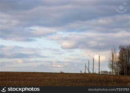 Plowed field and trees on the back, against a blue sky. Spring landscape with cornfield, wood and cloudy blue sky. Classic rural landscape in Latvia.