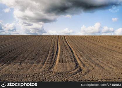 Plowed field with tracks under a blue sky with dark clouds in the spring