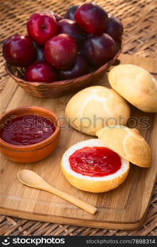 Plum jam spread on bun with plums in basket on wooden board photographed with natural light (Selective Focus, Focus on the front of the jam on the bun)