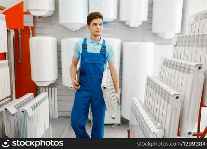 Plumber in uniform holds water heating radiator in plumbering store. Man buying sanitary engineering in shop. Plumber holds heating radiator, plumbering store