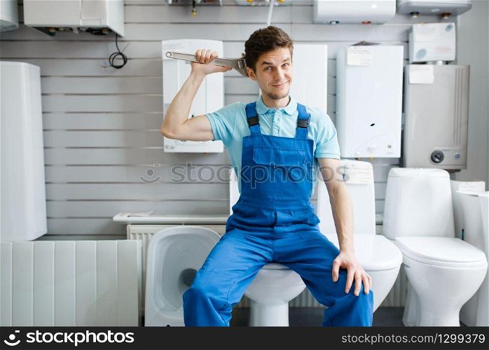 Plumber with pipe wrenches sitting on toilet at the showcase in plumbering store. Man buying sanitary engineering tools and equipment in shop. Plumber sitting on toilet in plumbering store