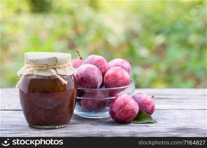 Plums in transparent bowl and jar of plum jam on wooden table. Natural green background. Plums in transparent bowl and jar of plum jam