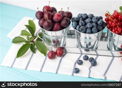 Plums, red currants and blueberries in small metal bucket on the wooden table.