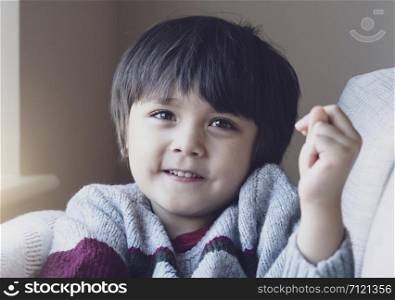 Poatrait of happy boy looking at camear with smiling face.Selective focus Adorable child with smiling face sitting on sofa, relaxing at home on weekend in Autumn or WInter, Warm and cozy scene in Pastel tone.