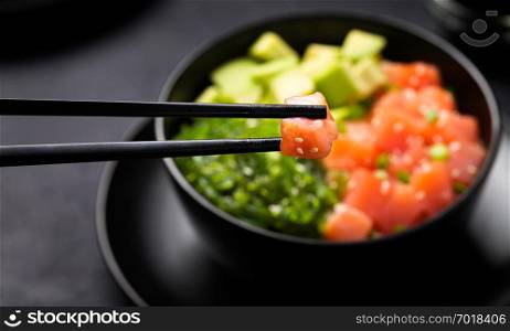 Poke with diced red fish, avocado and green seaweeds salad decorated with green onions and sesame seeds in black bowl on dark background with piece of salmon in chopsticks in foreground.. Poke with diced red fish, avocado and green seaweeds salad with piece of salmon in chopsticks in foreground.