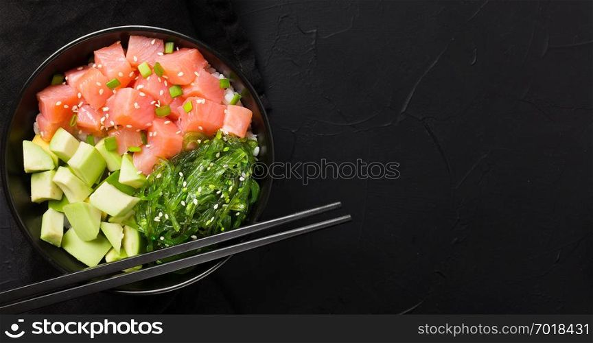 Poke with red fish, avocado and green seaweeds salad decorated with green onions and sesame seeds in black bowl with chopsticks on dark background - top view of seafood meal.. Poke with red fish, avocado and green seaweeds salad.