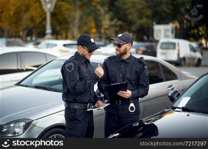 Police officers issue a ticket in the parking lot. Policemen in uniform protect the law, registration of an offense. Cops work on city street, order and justice control. Police officers issue a ticket in the parking lot