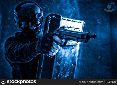Police quick reaction team, special operations tactical group member in mask, helmet and goggles, hiding behind ballistic shield, aiming with pistol in firefight with dangerous criminals or terrorists. SWAT team fighter aiming pistol from behind shield