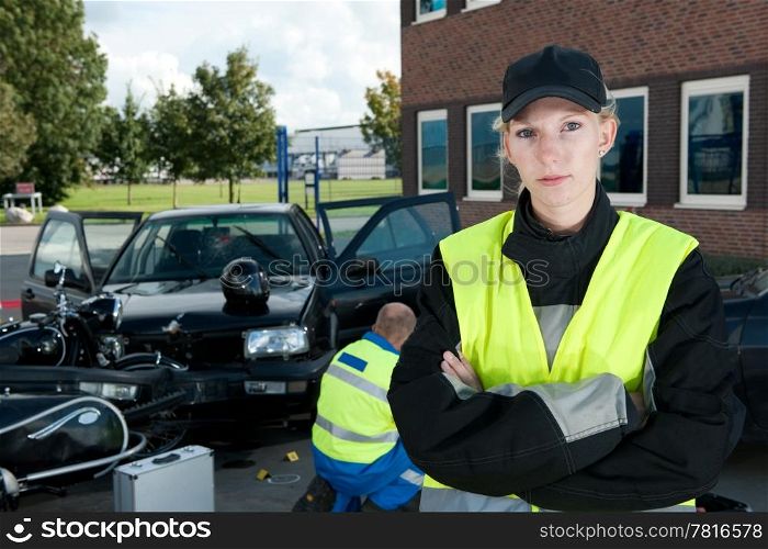 Police woman posing on the site of a car crash, with a paramedic working in the background