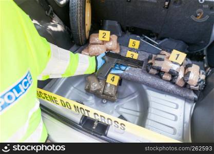 Policeman holding gun discovered in the trunk of a car, drug packages in the background