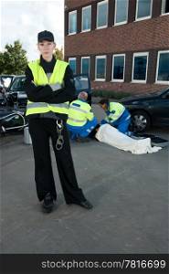 Polise woman posing for the camera at the site of a car crash, with two paramedics tending to an injured driver in the background