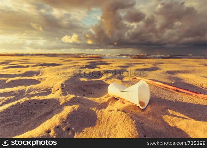 Polluted beach shore. Plastic disposable cup abandoned on sand, beautiful golden sunset in the background. Earth ecology. Plastic disposable cup on sea shore. Earth ecology