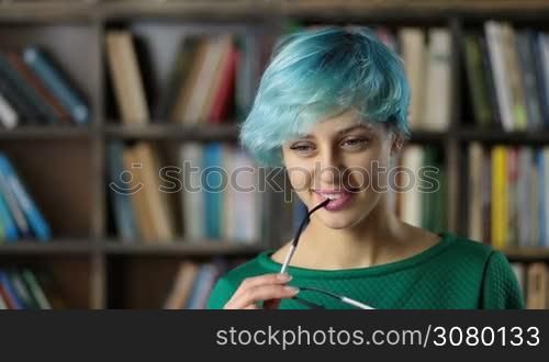 Pondering attractive female journalist with eyeglasses rim in her mouth thinking and generating article ideas for publication and smiling. Thoughtful gorgeous hipster girl pondering preparing areport for homework analyzing information at library.