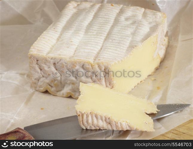 Pont l&rsquo;eveque cheese with its waxed paper wrapping resting on a board wit a slice cut from it.