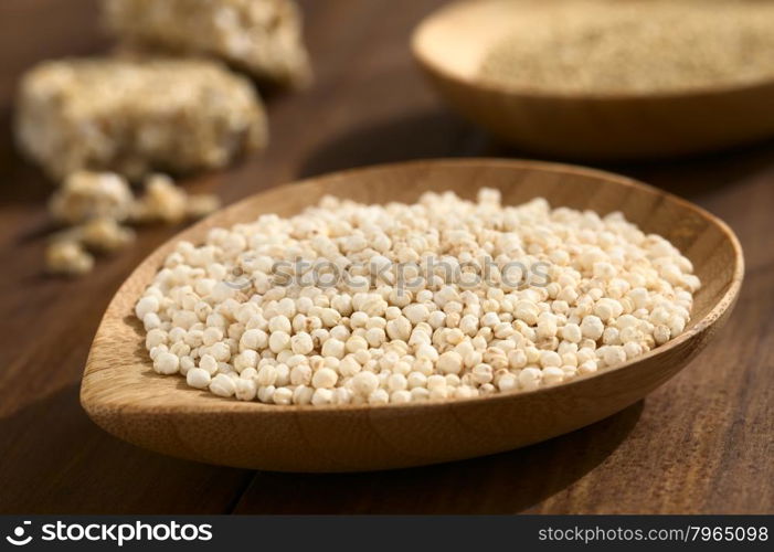 Popped white quinoa (lat. Chenopodium quinoa) cereal on small wooden plate, raw quinoa grains and quinoa cereal bar in the back, photographed on wood with natural light (Selective Focus, Focus one third into the quinoa cereal)