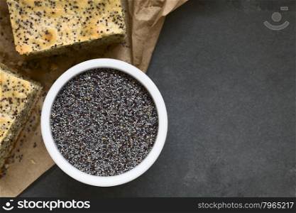 Poppy seeds in small bowl with poppy seed cake pieces on the side, photographed overhead on slate with natural light