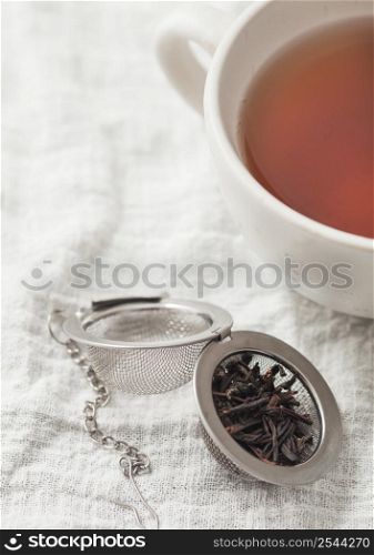 Porcelain cup with hot black tea and infuser with loose tea on white kitchen towel