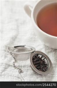 Porcelain cup with hot black tea and infuser with loose tea on white kitchen towel