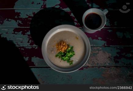 Pork congee or Rice porridge with minced pork sprinkled with deep-fried garlic and coriander in white bowl Served with cup of black coffee on old wooden table for new morning. Asia breakfast, Copy space, Top view, Selective focuse.