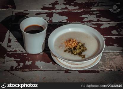 Pork congee or Rice porridge with minced pork sprinkled with deep-fried garlic and coriander in white bowl Served with cup of black coffee on old wooden table for new morning. Asia breakfast, Selective focuse.