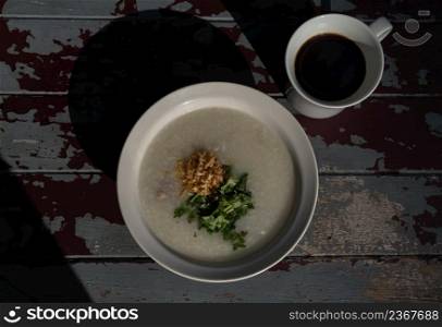 Pork congee or Rice porridge with minced pork sprinkled with deep-fried garlic and coriander in white bowl Served with cup of black coffee on old wooden table for new morning. Asia breakfast, Copy space, Top view, Selective focuse.