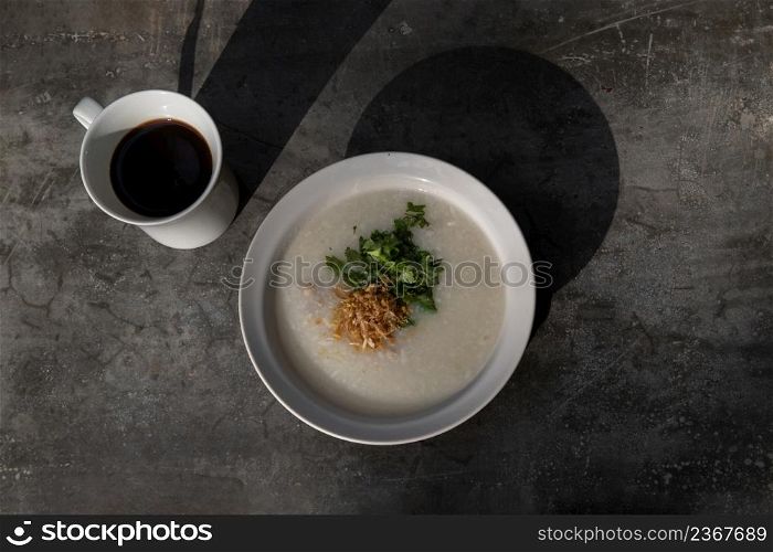 Pork congee or Rice porridge with minced pork sprinkled with deep-fried garlic and coriander in white bowl Served with cup of black coffee on stone table for new morning. Asia breakfast, Copy space, Top view, Selective focuse.