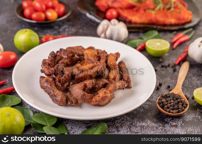 Pork cutlet in a white plate with pepper seeds, kaffir lime leaves, lemons, chilies, garlic on black cement.