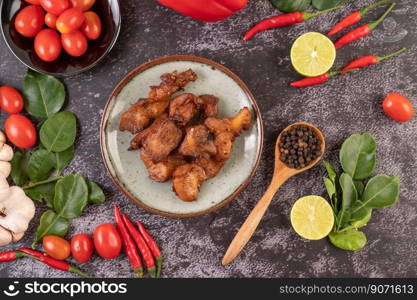 Pork cutlet in a white plate with pepper seeds, kaffir lime leaves, lemons, chilies, garlic on black cement.