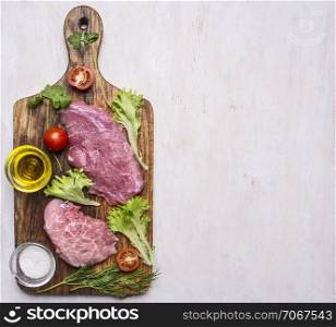 Pork steak with vegetables and herbs, meat knife and fork, on a cutting board with oil and seasonings border ,place for text on wooden rustic background top view close up