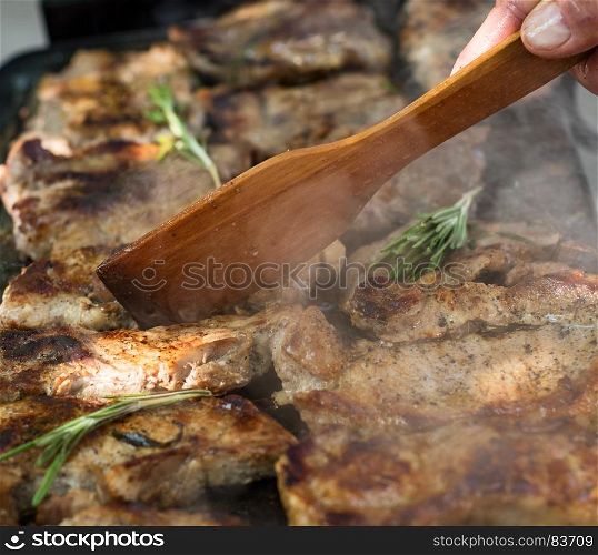 Pork steaks are roasted on a grill with rosemary branches, a human hand with a wooden spatula