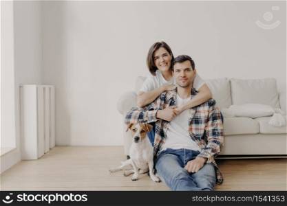 Portait of affectionate wife embraces husband who sits on floor near couch, their favourite pet poses at camera, make photo, being at home, buy new apartment, pose in living room with white walls