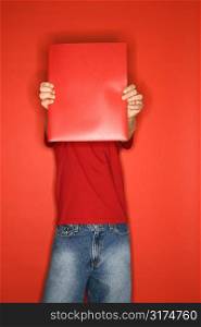 Portait of Caucasian boy covering his face with red folder standing against red background.