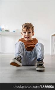 Portait of small caucasian boy cute little child kid sitting on the laminated wooden or vinyl floor at home in day alone smiling having fun playful