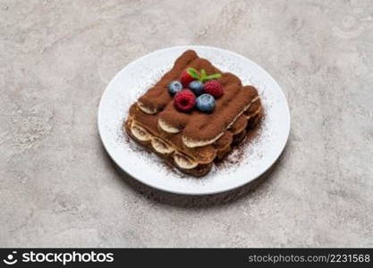 portion of Classic tiramisu dessert with raspberries and blueberries on grey concrete background or table. portion of Classic tiramisu dessert with raspberries and blueberries on grey concrete background