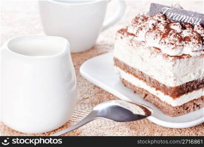 Portion of tiramisu dessert served on a white shaped plate and a cup of coffee with cream