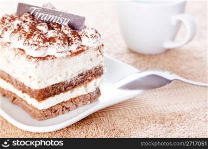 Portion of tiramisu dessert served on a white shaped plate and a cup of coffee